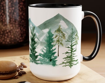 May include: White ceramic mug with a black rim and handle. The mug features a watercolor design of green trees and mountains.