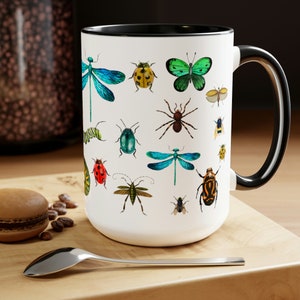 May include: White ceramic mug with a black rim and handle. The mug features a colorful design of various insects, including butterflies, dragonflies, beetles, and caterpillars.