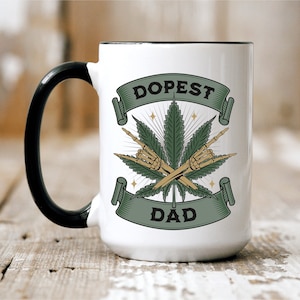 May include: White ceramic mug with a black handle. The mug has a green marijuana leaf design with two skeleton hands making the 'rock on' hand gesture. The text 'Dopest Dad' is printed on the mug.