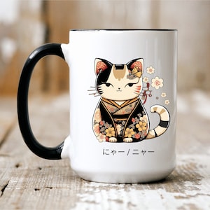 May include: White ceramic mug with a black handle. The mug features a cartoon illustration of a cat wearing a black and gold kimono with pink flowers. The cat has black, white, and orange fur. The text "にゃー/ニャー" is printed on the bottom of the mug.