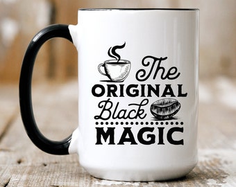 May include: White ceramic coffee mug with a black handle and black rim. The mug features a black and white illustration of a coffee cup with steam rising from it, a coffee bean, and the text "The Original Black Magic".