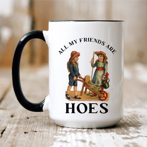 May include: White ceramic mug with a black handle. The mug has a black rim and features a vintage illustration of two children with a wheelbarrow. The text on the mug reads "All my friends are hoes".