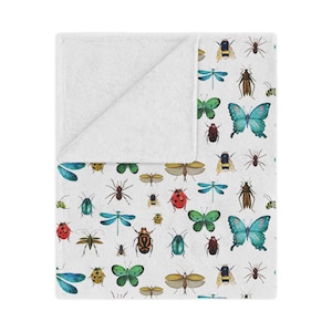 May include: White fleece throw blanket with a colorful pattern of various insects and butterflies. The pattern includes ladybugs, dragonflies, beetles, and moths.