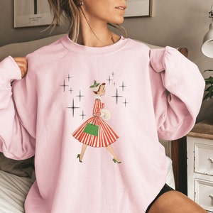 May include: Pink sweatshirt featuring a vintage illustration of a woman in a red and white striped dress carrying a green shopping bag and a white package. The illustration is surrounded by black stars.