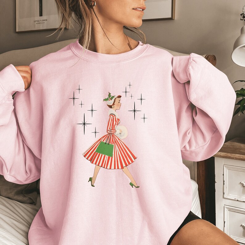 May include: Pink sweatshirt featuring a vintage illustration of a woman in a red and white striped dress carrying a green shopping bag and a white package. The illustration is surrounded by black stars.