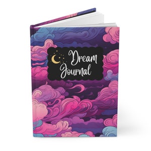 May include: A purple and pink dream journal with a black and white cover featuring a crescent moon and stars. The text "Dream Journal" is written in white cursive lettering.