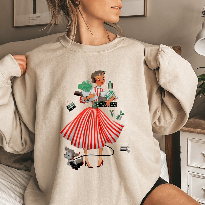 May include: A beige sweatshirt with a vintage illustration of a woman in a red and white striped dress carrying presents and walking a small dog.