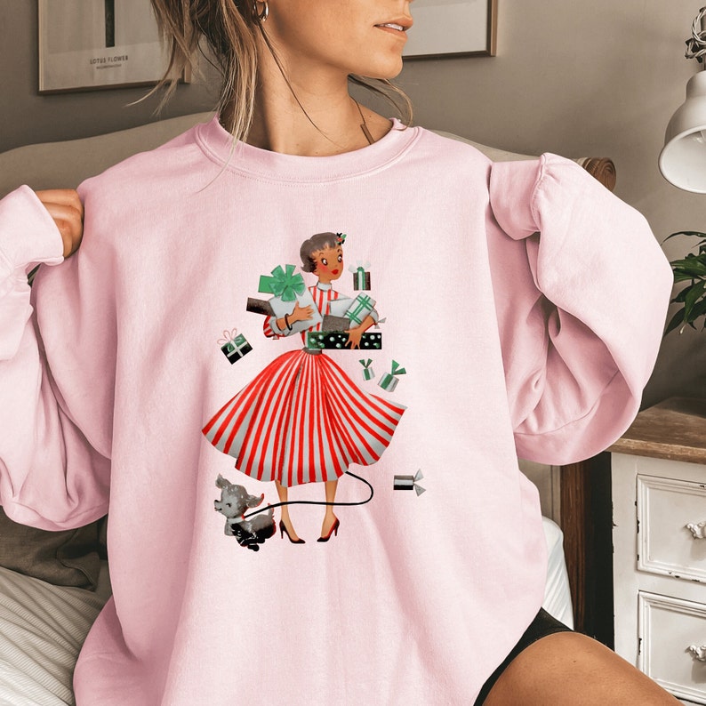 May include: Pink sweatshirt with a graphic print of a woman in a red and white striped dress holding presents. The woman is wearing a red bow in her hair and is surrounded by presents. A small dog is walking behind her.