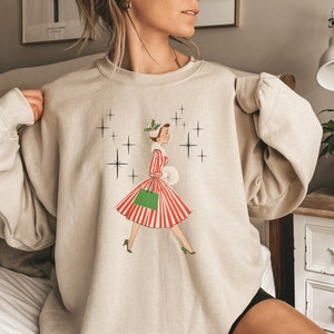 May include: A beige sweatshirt featuring a vintage illustration of a woman in a red and white striped dress carrying a green shopping bag. The illustration is surrounded by white stars.