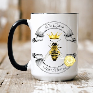 May include: White ceramic mug with a black handle. The mug features a graphic of a bee wearing a crown and the text "The Queen Has Retired".