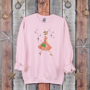 May include: Pink sweatshirt featuring a vintage illustration of a woman in a red and white striped dress and green handbag walking through a snowy scene with sparkling stars.