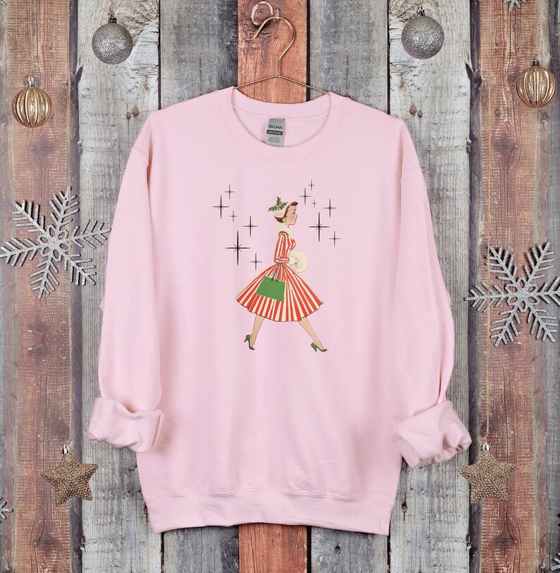 May include: Pink sweatshirt featuring a vintage illustration of a woman in a red and white striped dress and green handbag walking through a snowy scene with sparkling stars.