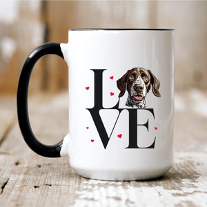 May include: White ceramic coffee mug with a black handle. The mug features a black and white illustration of a German Shorthaired Pointer dog with the word "LOVE" in black letters and red hearts around the letters.