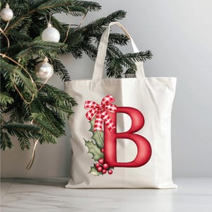 May include: A cream-colored tote bag featuring a large red letter "B" decorated with a red and white gingham bow, holly leaves, and red berries. The bag has a simple design with a rectangular shape and handles. A Christmas tree with ornaments is in the background.