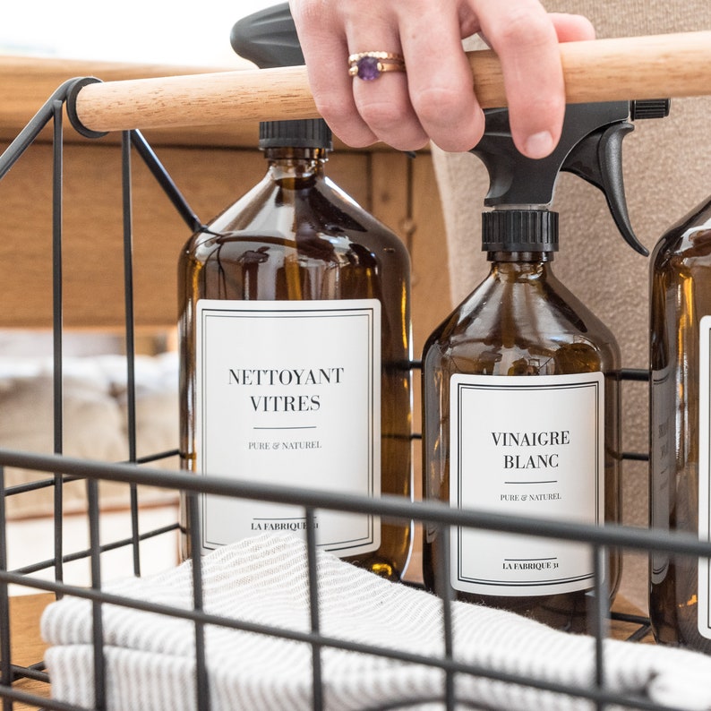 May include: Three amber glass bottles with black spray tops and white labels. The labels read "Nettoyant Vitres Pure & Naturel La Fabrique" and "Vinaigre Blanc Pure & Naturel La Fabrique". The bottles are in a black wire basket.