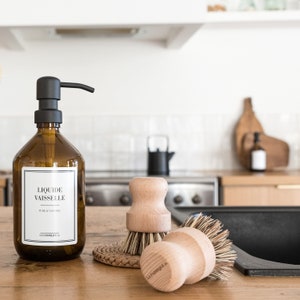 May include: A brown glass bottle with a black pump dispenser labeled "Liquide vaisselle" and "La Fabrique 19". The bottle is sitting on a wooden countertop next to two wooden dish brushes with natural bristles.