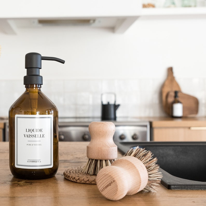 May include: A brown glass bottle with a black pump dispenser labeled "Liquide vaisselle" and "La Fabrique 19". The bottle is sitting on a wooden countertop next to two wooden dish brushes with natural bristles.