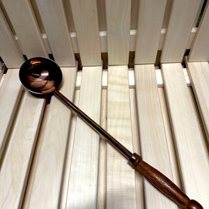 May include: A copper-coloured ladle with a long, slender handle and a wooden grip. The ladle's bowl is round and reflective. The handle is dark brown and tapers to a rounded end. The ladle is resting on a light-coloured wooden surface.