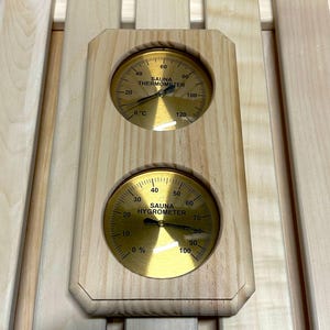 Puede incluir: Un termómetro e higrómetro de sauna de madera. El termómetro muestra temperaturas en grados Celsius, de 0 a 120 grados. El higrómetro indica los niveles de humedad del 0% al 100%. Ambos medidores tienen una esfera dorada.