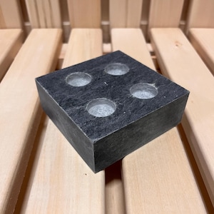 Puede incluir: Una piedra cuadrada de pizarra negra con cuatro hendiduras redondas. La piedra está sobre una superficie de madera.