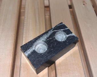 May include: A black marble candle holder with two circular openings. The holder is sitting on a wooden surface.