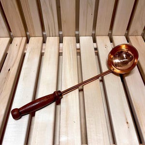 May include: A copper ladle with a wooden handle. The ladle is resting on a wooden surface.