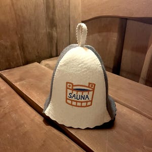 Könnte beinhalten: Eine weiße und graue Filz-Saunamütze mit einem blauen und orangefarbenen gestickten Saunaeimer-Logo.