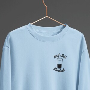 Peut inclure: Un sweat-shirt bleu clair avec un graphique d'un verre à moitié plein et le texte "Half-Full Mindset".