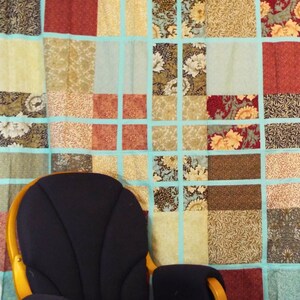 May include: A black rocking chair with wooden arms sits in front of a wall covered in a patchwork quilt. The quilt is made of various fabrics in shades of brown, beige, red, and blue.