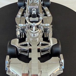 May include: A detailed, chrome-colored model of a Formula 1 race car. The model features intricate details of the engine, chassis, and wheels. The car is positioned on a black surface, showcasing its sleek design.