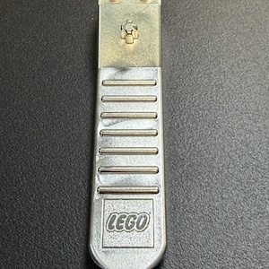 May include: A silver-colored LEGO brick separator tool. The tool has the LEGO logo at the bottom and two LEGO logos at the top. The tool has a series of horizontal lines and a small cross-shaped design.