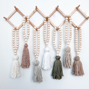 May include: Wooden bead garland with decorative tassels in various colors, including white, beige, and green. The garland is suspended from a natural wood wall-mounted rack. This home decor item adds a touch of bohemian style.