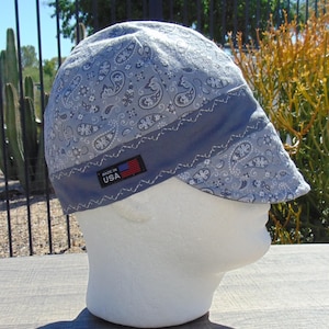 May include: A light blue paisley print welding cap with a dark blue band and a small "MADE IN USA" tag. The cap is displayed on a white mannequin head. The paisley pattern includes white and dark blue floral and swirl designs.
