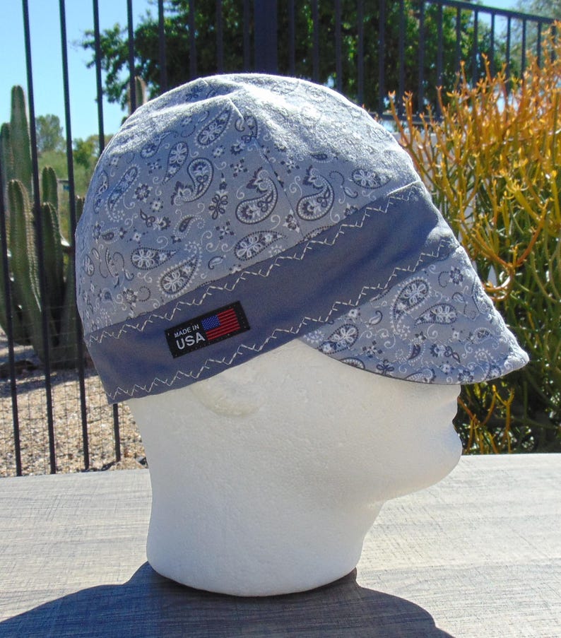 May include: A light blue paisley print welding cap with a dark blue band and a small "MADE IN USA" tag. The cap is displayed on a white mannequin head. The paisley pattern includes white and dark blue floral and swirl designs.