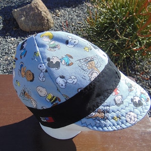 May include: A blue and black baseball cap with a Peanuts cartoon print. The cap has a black brim and a black band around the crown. The band has a small American flag patch on it. The cap is made in the USA.