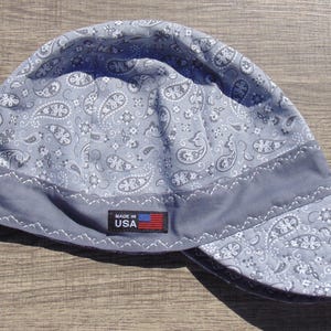 May include: A gray welding cap with a paisley pattern and a dark gray band. The cap has a small tag that reads "MADE IN USA" with a US flag. The cap is made of fabric.