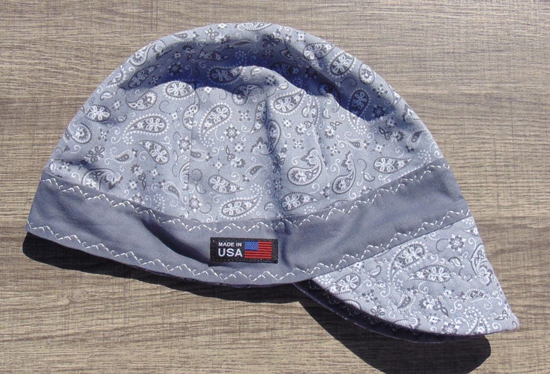 May include: A gray welding cap with a paisley pattern and a dark gray band. The cap has a small tag that reads "MADE IN USA" with a US flag. The cap is made of fabric.