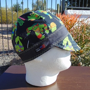 May include: A black cap featuring a colorful pattern of Teenage Mutant Ninja Turtles characters. The cap has a dark brim with zigzag stitching and a small "Made in USA" tag with the American flag.