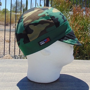 May include: A welding cap featuring a green, brown, and black camouflage pattern with a solid green band. The cap includes a small black rectangular tag on the side with the text MADE IN USA and a graphic of the American flag in red, white, and blue.