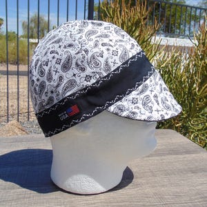 May include: A white and black paisley print welding cap with a black band and white stitching. The cap has a small tag with the text "MADE IN USA" and a small American flag.
