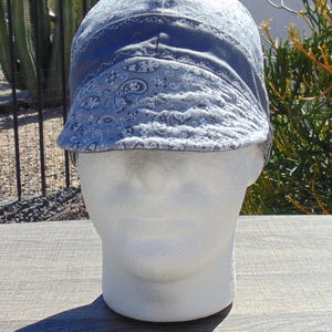 May include: A blue welding cap with a paisley pattern. The cap is on a white mannequin head. The cap has a brim and is made of fabric. The background includes plants and a wooden surface.