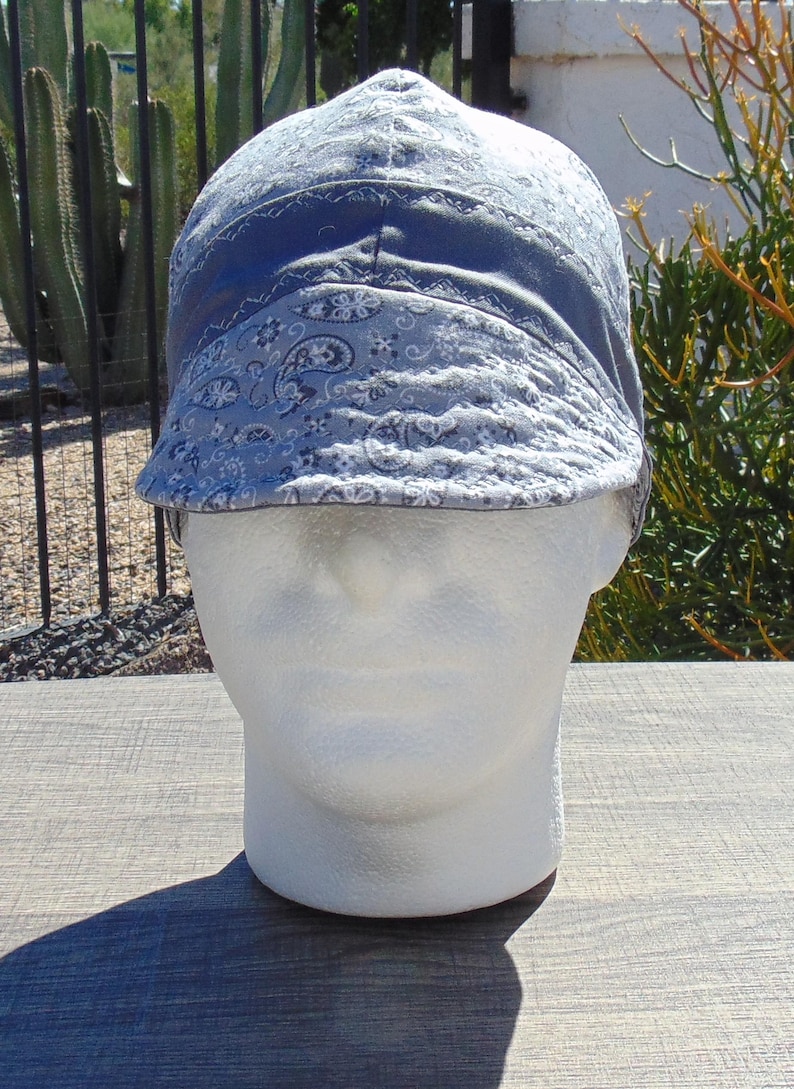 May include: A blue welding cap with a paisley pattern. The cap is on a white mannequin head. The cap has a brim and is made of fabric. The background includes plants and a wooden surface.