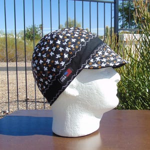 May include: A black and white patterned welding cap with a black brim and a white zig-zag stitch. The cap has a pattern of white and orange shapes. The label reads "Made in USA".