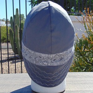 May include: A blue welding cap with a gray paisley band and white stitching. The cap is made of a durable fabric and is designed to protect the wearer from sparks and heat. The cap is displayed on a white mannequin head.