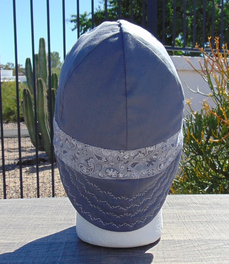 May include: A blue welding cap with a gray paisley band and white stitching. The cap is made of a durable fabric and is designed to protect the wearer from sparks and heat. The cap is displayed on a white mannequin head.