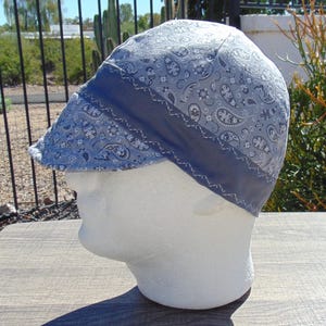 May include: A light blue paisley print welding cap with a dark blue band and white stitching. The cap is displayed on a white mannequin head, showcasing its design and fit. The cap is designed to protect the head from sparks and heat.