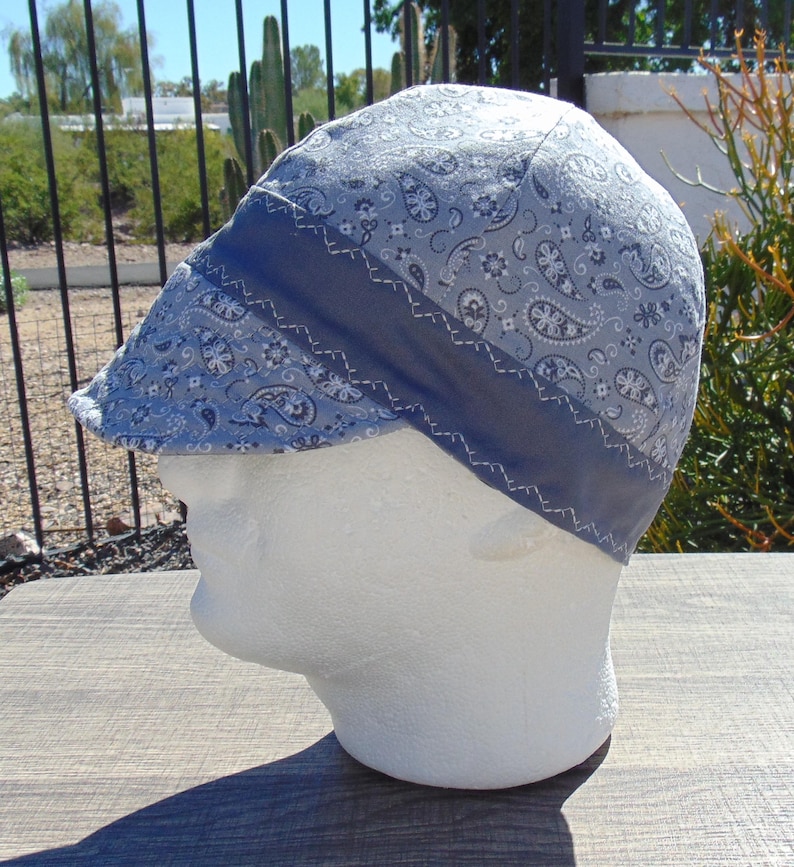 May include: A light blue paisley print welding cap with a dark blue band and white stitching. The cap is displayed on a white mannequin head, showcasing its design and fit. The cap is designed to protect the head from sparks and heat.