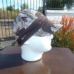 May include: A brown and tan camouflage welding cap with a brown brim and a black label that says "Made in USA".