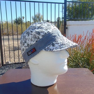 May include: A white and black paisley patterned welding cap with a gray brim and a small "Made in USA" tag. The cap is displayed on a white mannequin head, showcasing its design and construction.