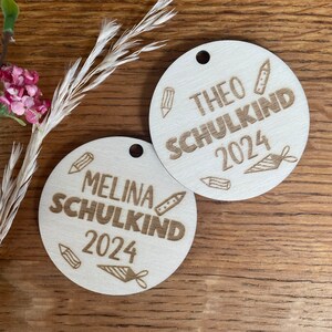 Op de afbeelding: Twee ronde houten labels met een gat aan de bovenkant. Eén label leest "THEO SCHULKIND 2024" en de andere "MELINA SCHULKIND 2024". Beide hebben potlood- en schooltas illustraties. Op een houten oppervlak.